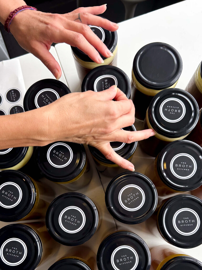 Hands meticulously placing labels on jars with black lids, marked as "Organic Grass-Fed Beef Bone Broth 720g," home to the rich offerings of The Broth Kitchen.