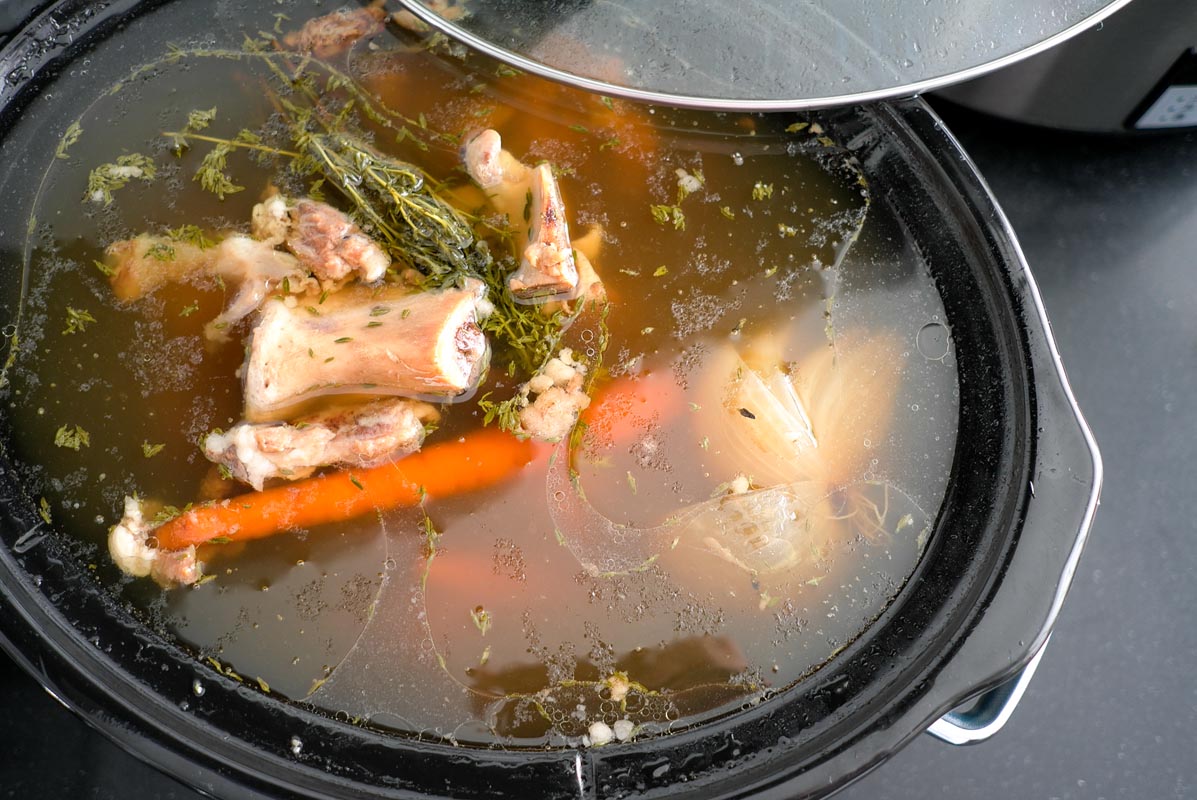 A slow cooker filled with organic grass-fed beef bone broth ingredients, including bones, carrots, onions, and herbs submerged in liquid, with the lid partially covering the pot.